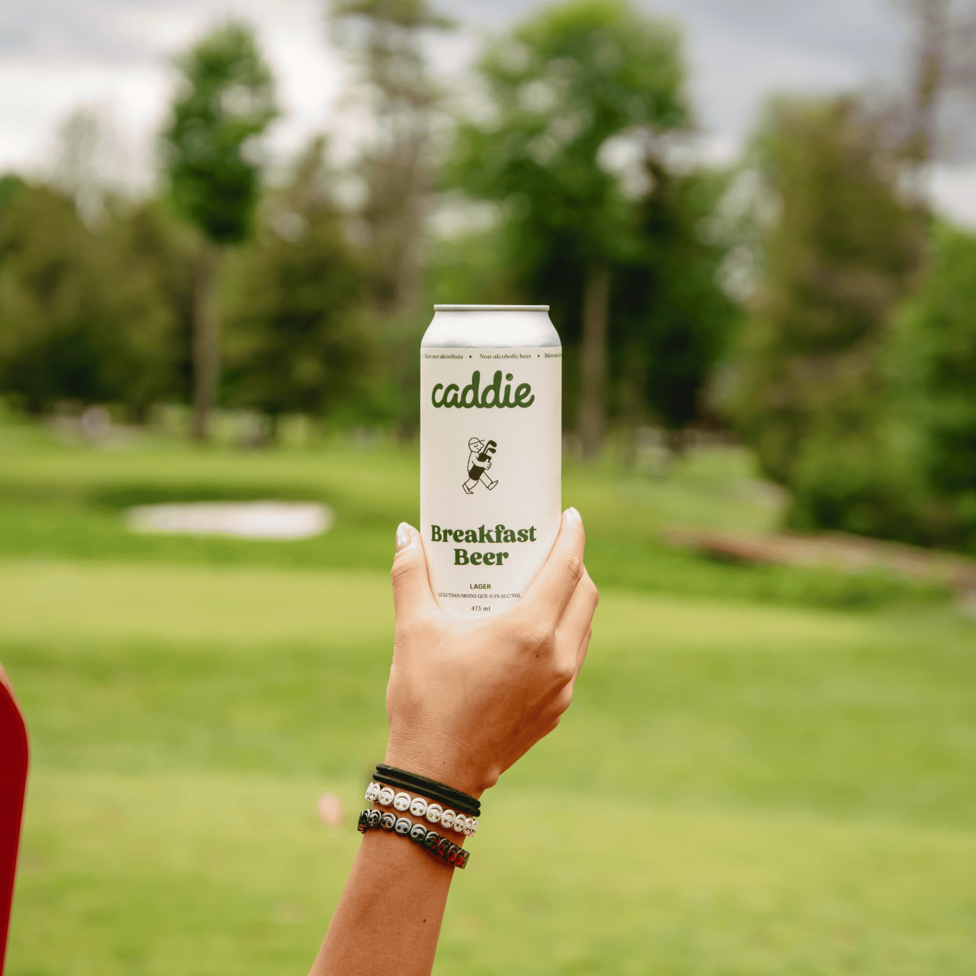A hand raising a Caddie Breakfast Beer tallboy against a fairway with a bunker and trees.