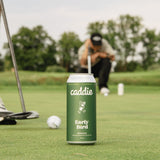 A green can of caddie Early Bird beer sits on a golf green in the foreground, with a golfer crouching in the blurred background on the putting green.